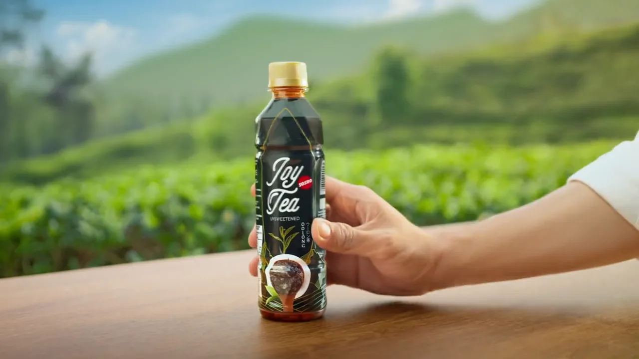 Joy Tea key visual: a hand holding a bottle of unsweetened Joy Tea on a wooden table before tea plantation hills
